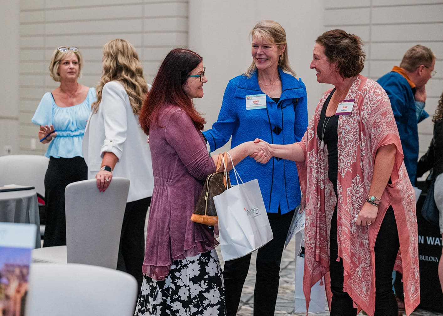 A group of people interact at a Retreats Resources Roadshow. Two women in the foreground shake hands while holding gift bags. Other attendees are seen in the background.