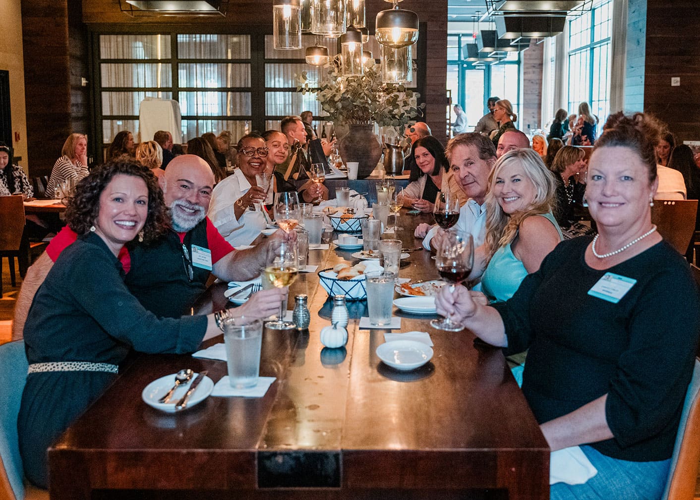A group of people sit around a long table in a restaurant, smiling and raising glasses, with others dining in the background.