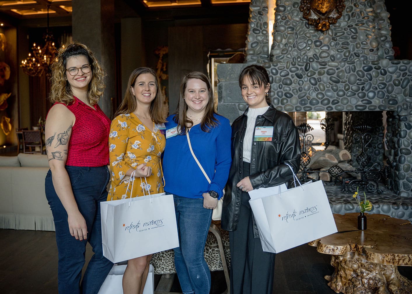 Four women stand indoors holding white gift bags, smiling at the camera in front of a decorative stone fireplace.