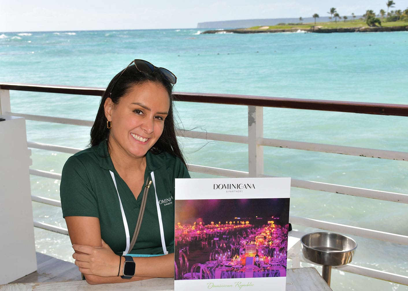 A woman sits at a table by the ocean, smiling at the camera with a Dominicana sign in front of her.