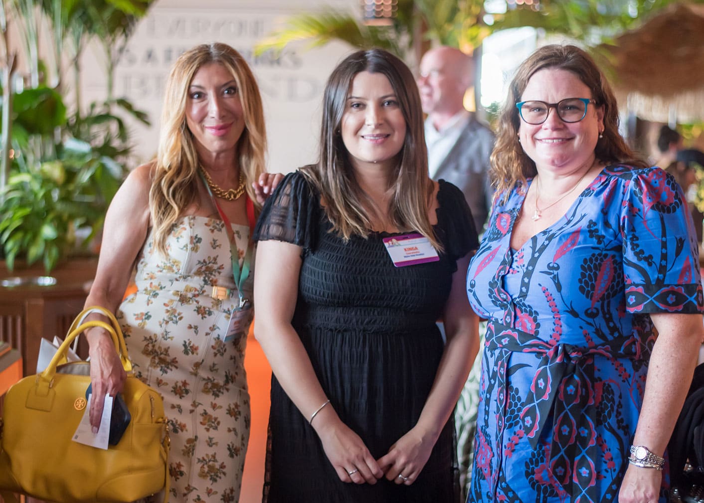 Three women stand together indoors at an event, each smiling at the camera. One holds a yellow handbag and all wear event badges. Lush green plants are visible in the background.