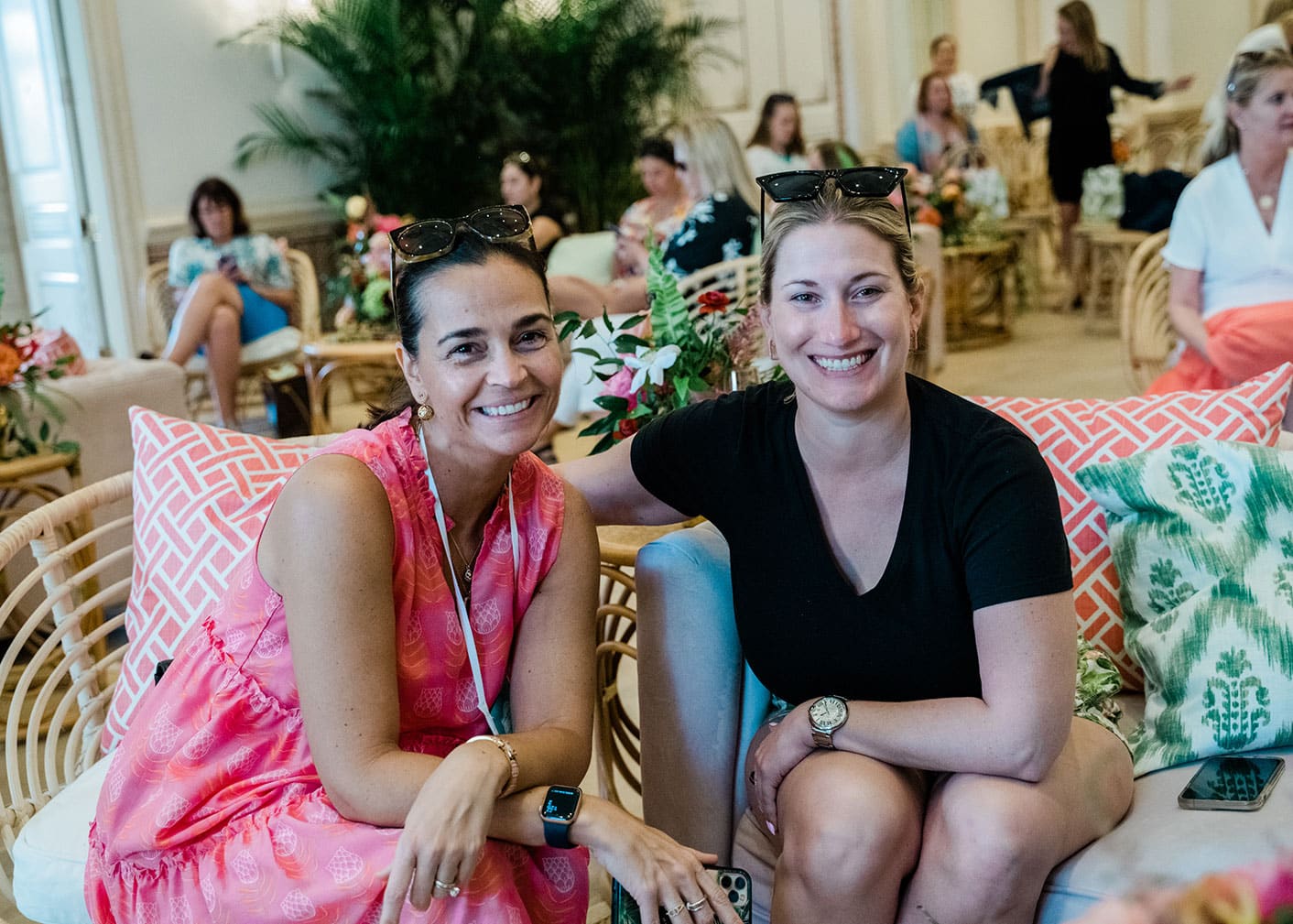 Two women sit smiling on a sofa in a bright, decorated room with other people seated in the background.