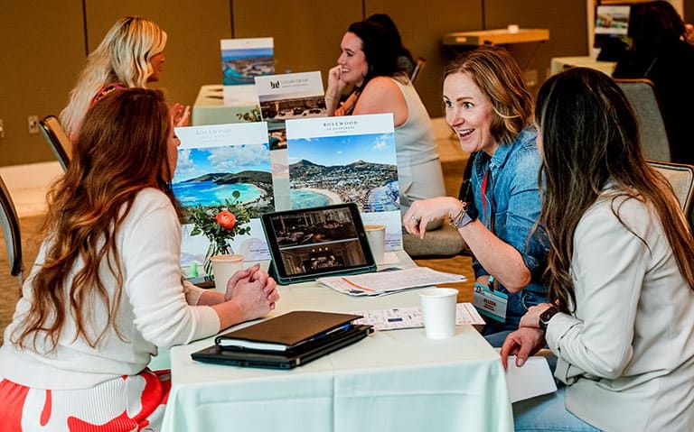 During a Retreats Resources Roadshow B2B Marketplace, three women sit at a table in discussion with travel brochures and a laptop, while others meet at tables in the background.