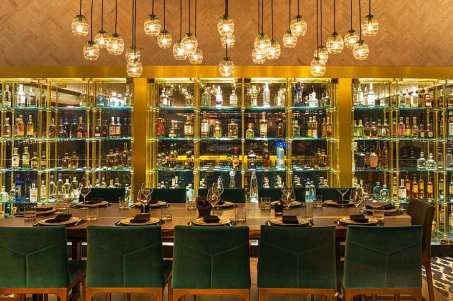 The Mexican, Dallas, TX – A dining table set for eight is in front of a large, illuminated wall of liquor bottles, with hanging glass pendant lights overhead.