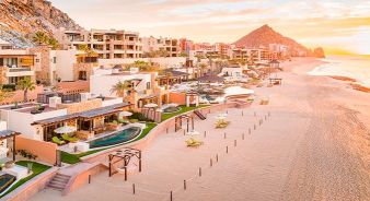 Waldorf Astoria Los Cabos Pedregal beachfront resort with modern villas, pools, loungers, and cabanas along a sandy shoreline at sunset; ocean waves on the right, rocky hills in the background.