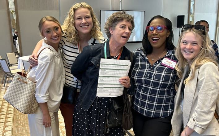 Five women stand together indoors, smiling at the camera at a Retreats Resources showcase in Park City, Utah. One woman in the center holds sheets of paper with graphs or tables.