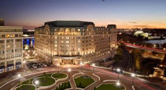 A large, well-lit Salamander DC hotel building stands at a curved intersection near a river at dusk, with city lights, trees, and cars visible.
