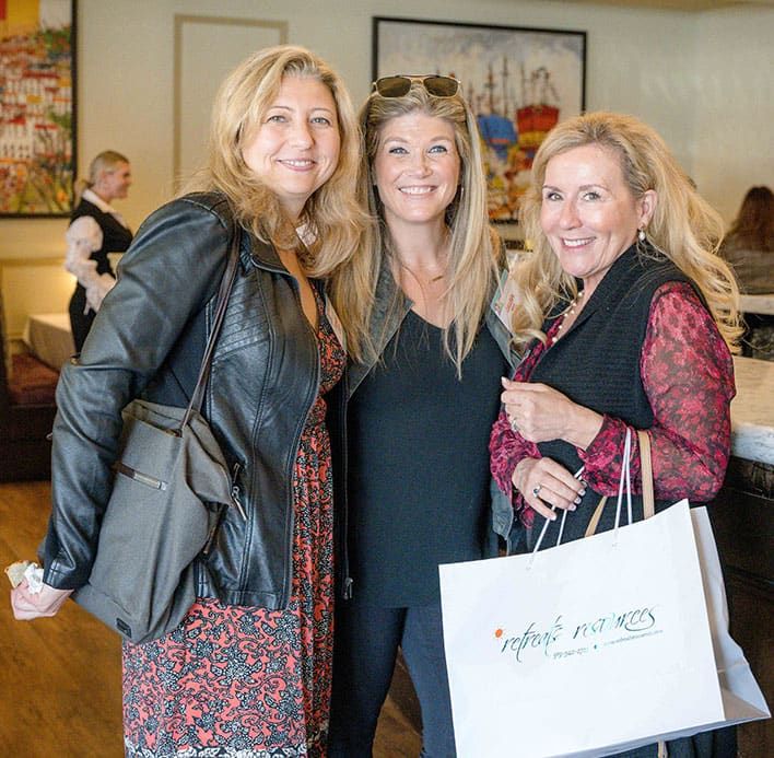 Three women stand indoors, smiling at the camera. One holds a large white Retreats Resources goodie bag. Artworks are visible on the wall behind them.