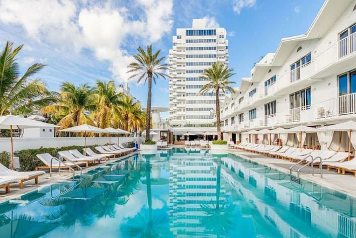The Shelborne by Proper outdoor swimming pool with lounge chairs, palm trees, and cabanas, set against a backdrop of modern white buildings under a partly cloudy sky.