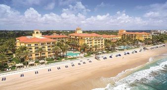 Eau Palm Beach Resort & Spa - Manalapan, FL - a large beachfront resort with yellow buildings, red roofs, palm trees, multiple pools, and rows of blue lounge chairs on a sandy beach by the ocean under a partly cloudy sky.