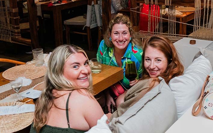 Three smiling women are seated at a table in a cozy indoor setting, enjoying drinks at a Retreats Resources roadshow..