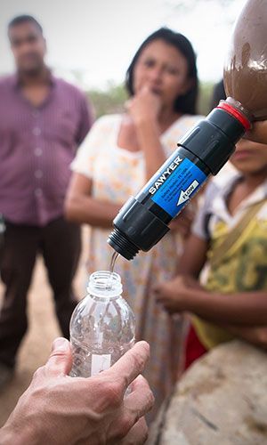 A person fills a plastic bottle with water using a Sawyer water filter, while three people watch in the background.