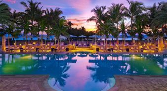 St. Regis Bal Harbour resort pool surrounded by lounge chairs and palm trees at sunset, with colorful sky reflecting on the water.