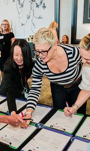 Three smiling women writing their competing bids in a Retreats Reources silent auction. The room is adorned with floral wall art.