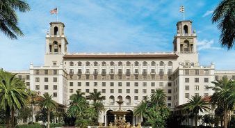 The large, historic The Breakers hotel in Palm Beach FL, with two towers, palm trees, an American flag, and a central fountain in front of the entrance.