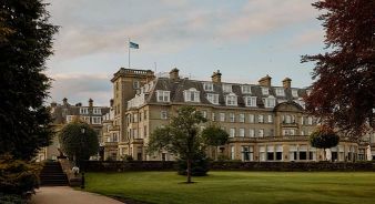 The Gleneagles Hotel, Perthshire, Scotland - a large, historic hotel building with multiple windows and chimneys, surrounded by manicured lawns and trees under a partly cloudy sky.