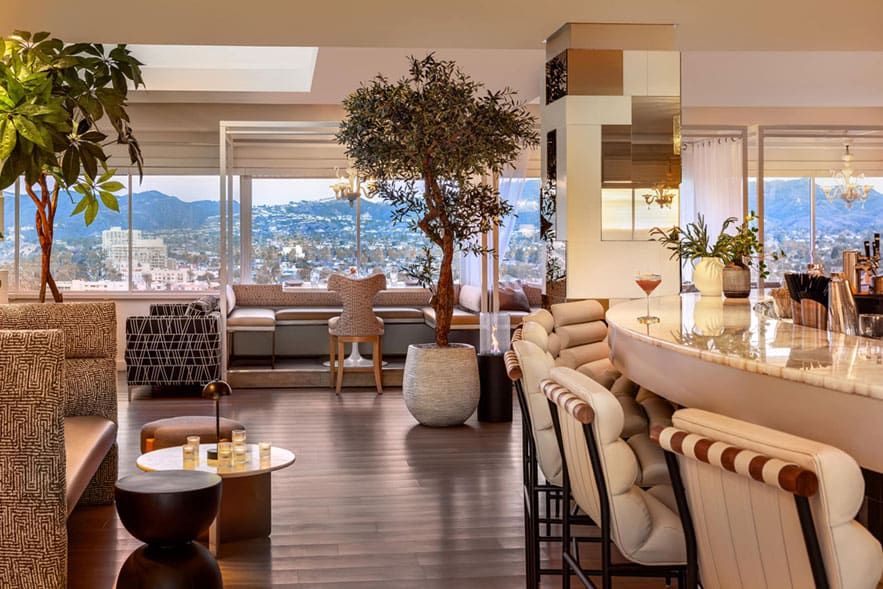 A modern lounge at The Huntley Hotel, with a curved bar, cushioned stools, indoor plants, and large windows offering a city and mountain view.