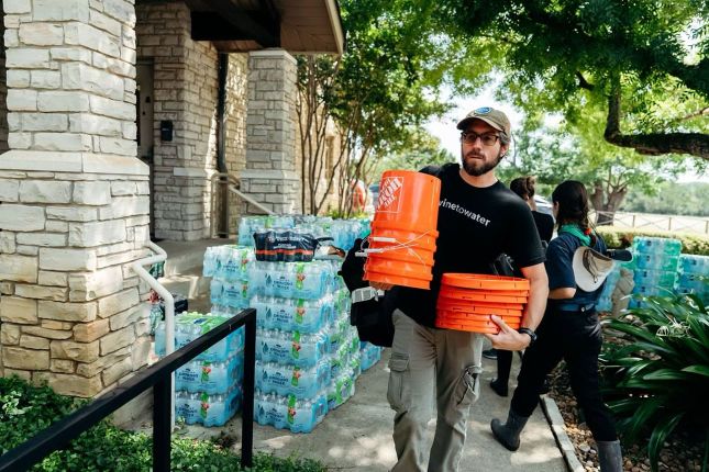 A man carries several orange buckets and supplies past stacks of bottled water outside a building, while others work in the background.