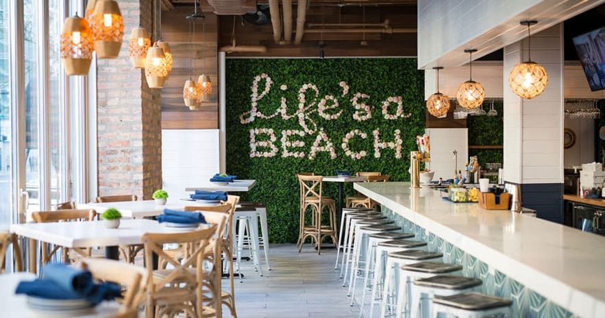 The Hampton Social River North in Chicago, IL. Bright cafe interior with wooden tables, white bar stools, hanging pendant lights, and a green wall featuring the words "Life's a BEACH" in white script.