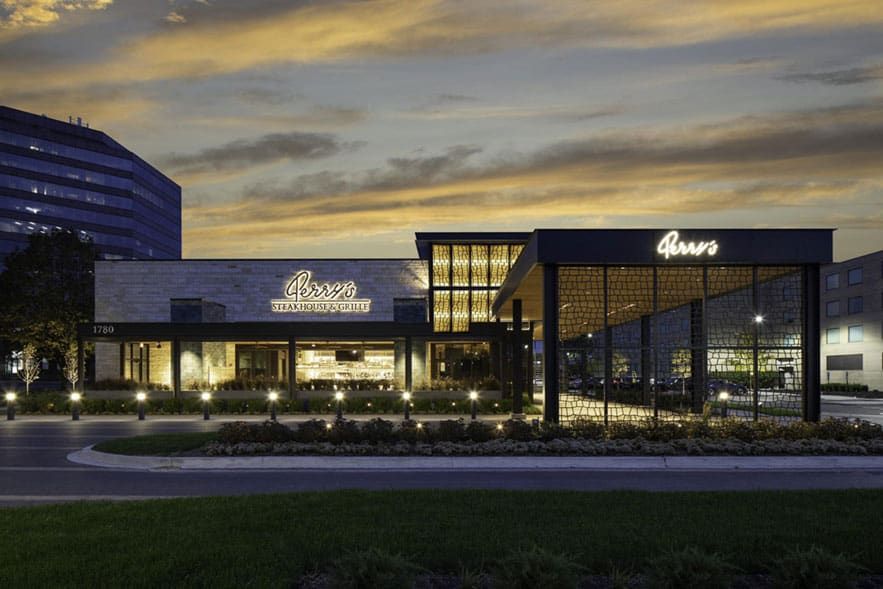 Modern steakhouse and grill restaurant exterior at dusk, illuminated signage reading “Perry’s Steakhouse & Grill” with large windows and landscaped entrance.