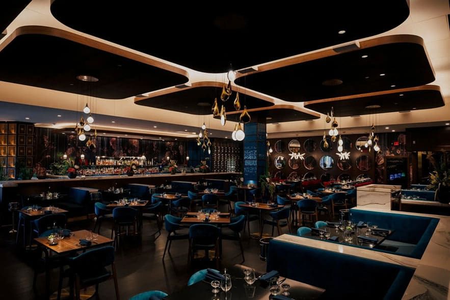 Boketto steakhouse in St Louis Park, MN. Modern restaurant interior with dark walls, blue velvet chairs, wooden tables, and artistic hanging light fixtures, featuring a visible bar area in the background.
