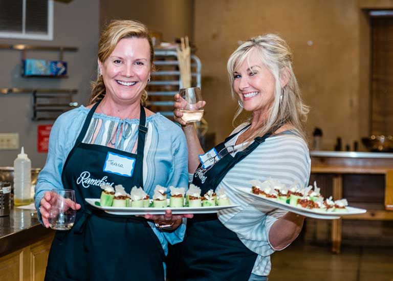 Two women wearing aprons hold trays of appetizers and drinks, smiling in a kitchen setting.