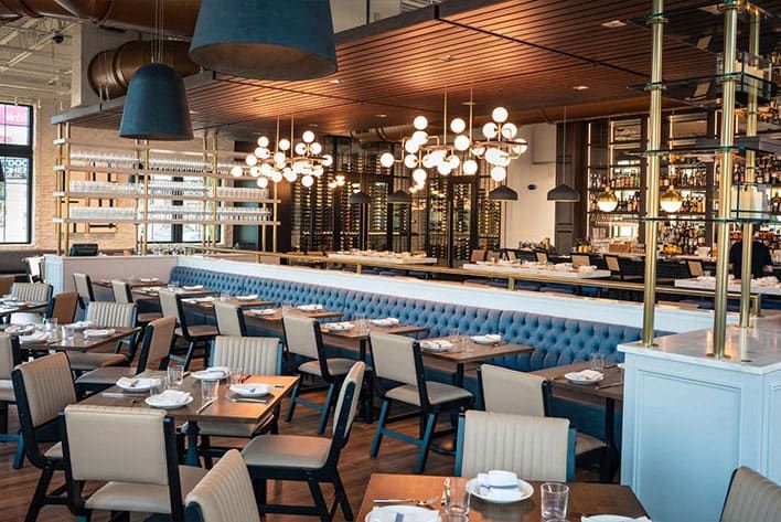 Olivia Tampa - Modern restaurant interior with wooden tables, upholstered chairs, blue banquette seating, and decorative pendant lighting above a bar area.