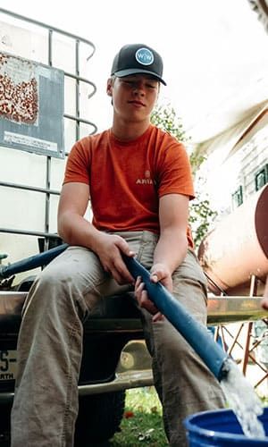 A person wearing a cap and orange shirt sits on a truck tailgate, holding a blue hose with water flowing into a blue bucket.