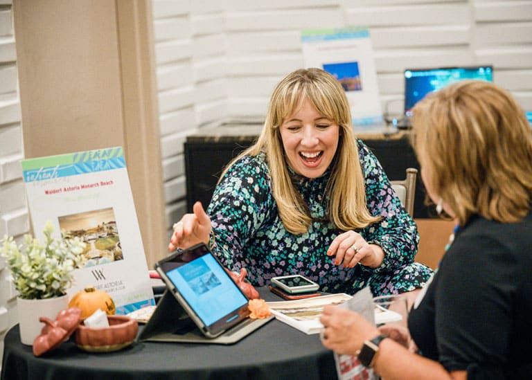 Two women sit at a table with brochures, a tablet, and promotional materials, engaged in a lively conversation during a Retreats Resources’ luxury hospitality showcase.