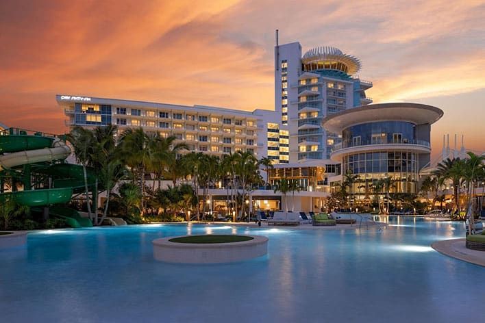  Pier Sixty-Six - A modern resort with palm trees, water slides, and a large pool is shown at sunset, with the buildings lit up against an orange sky.