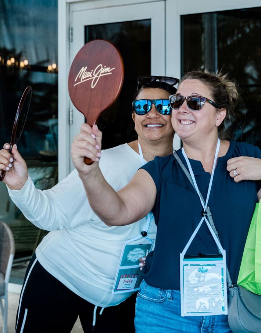 Two women wearing sunglasses smile and pose for a selfie while holding Maui Jim branded paddles, standing in front of glass doors at an outdoor event.