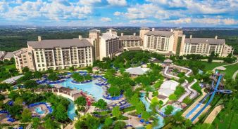Aerial view of the large JW Marriott San Antonio Hill Country Resort & Spa resort complex featuring multiple hotel buildings, landscaped gardens, swimming pools, and winding water slides under a partly cloudy sky.