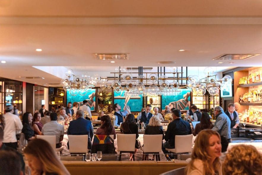 A busy Steak 48 Del Mar restaurant bar scene with people dining and socializing under modern lighting, with shelves of bottles and decorative artwork in the background.