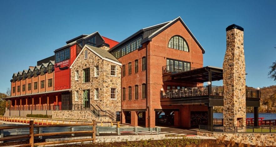 River House at Odette’s - A large riverside building with stone and brick exterior, multiple floors, large windows, outdoor terrace, and a tall stone chimney, set against a clear blue sky.