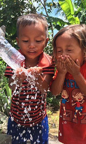 Two young children stand outside with trees behind them, catching and playing with water flowing from a faucet, smiling and laughing.