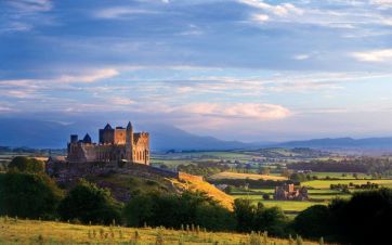 Stone castle complex situated on a green hilltop overlooking a rural landscape with distant mountains under a partly cloudy sky at sunset.