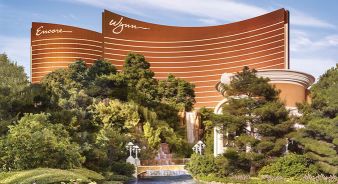 The Wynn and Encore Las Vegas hotels are shown with their curved bronze facades, surrounded by trees, a waterfall, and landscaped gardens in front.