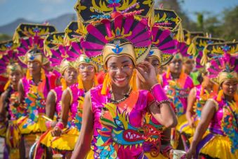 A group of people in colorful, elaborate costumes and headdresses smile during a festive outdoor parade.