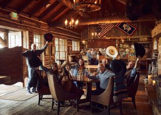 A group of people sit around a table in a rustic log cabin, raising cowboy hats and smiling, with wooden beams, western decor, and flags in the background.