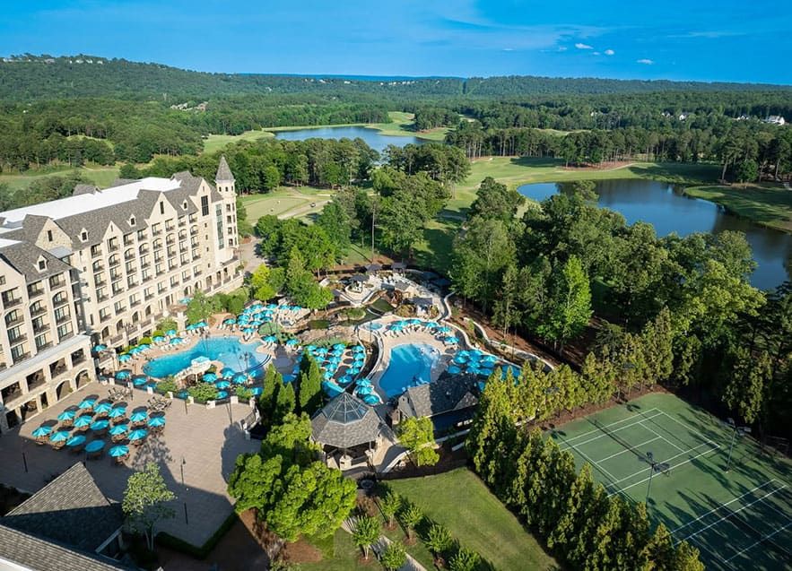 Aerial view of the large Renaissance Ross Bridge resort with outdoor swimming pools, tennis courts, and surrounding forested landscape with lakes and hills.