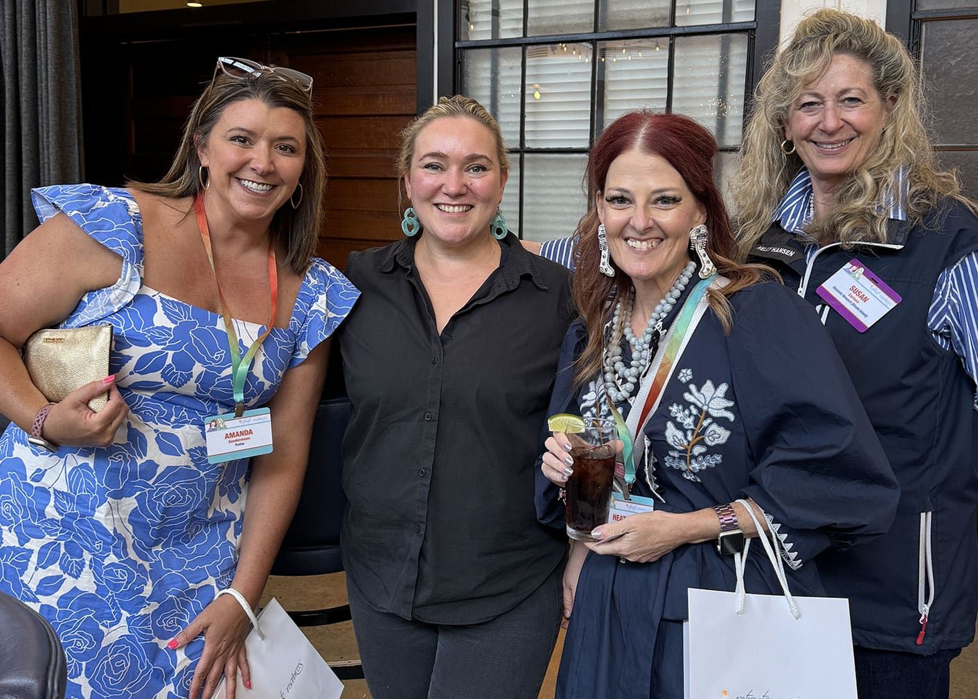 Four women stand together indoors, smiling at the camera. Three wear name tags and hold gift bags; one holds a drink. They are dressed in casual and semi-formal attire.
