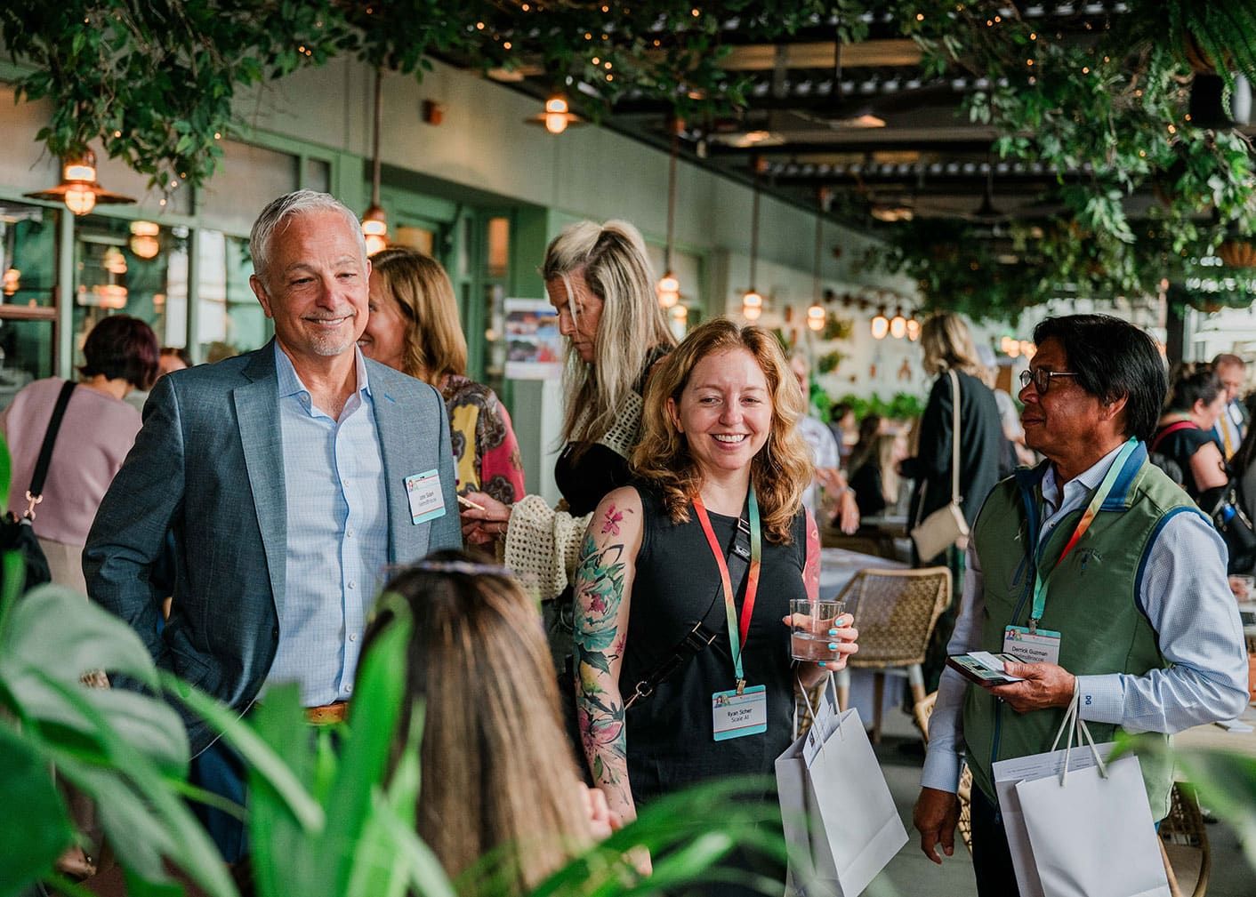 A group of people wearing name badges and event lanyards converse and smile at an indoor gathering with greenery and ambient lighting.