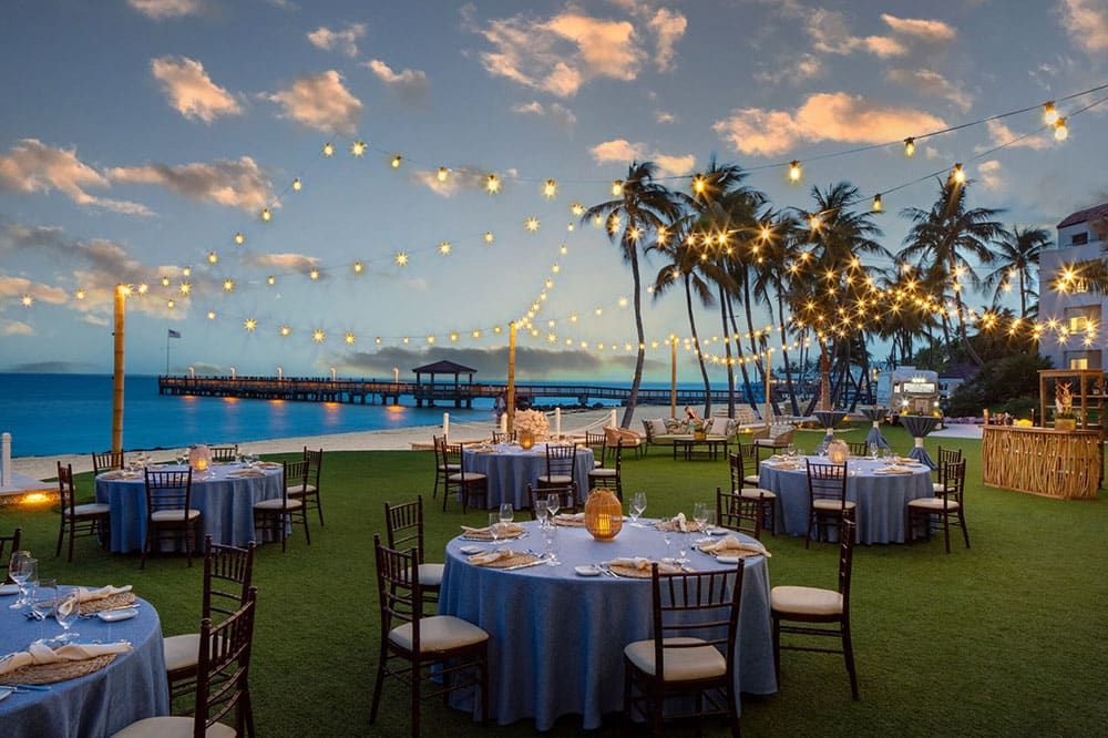 Round tables with blue tablecloths and wooden chairs are set up on a lawn by the ocean, with string lights overhead and palm trees in the background at sunset.