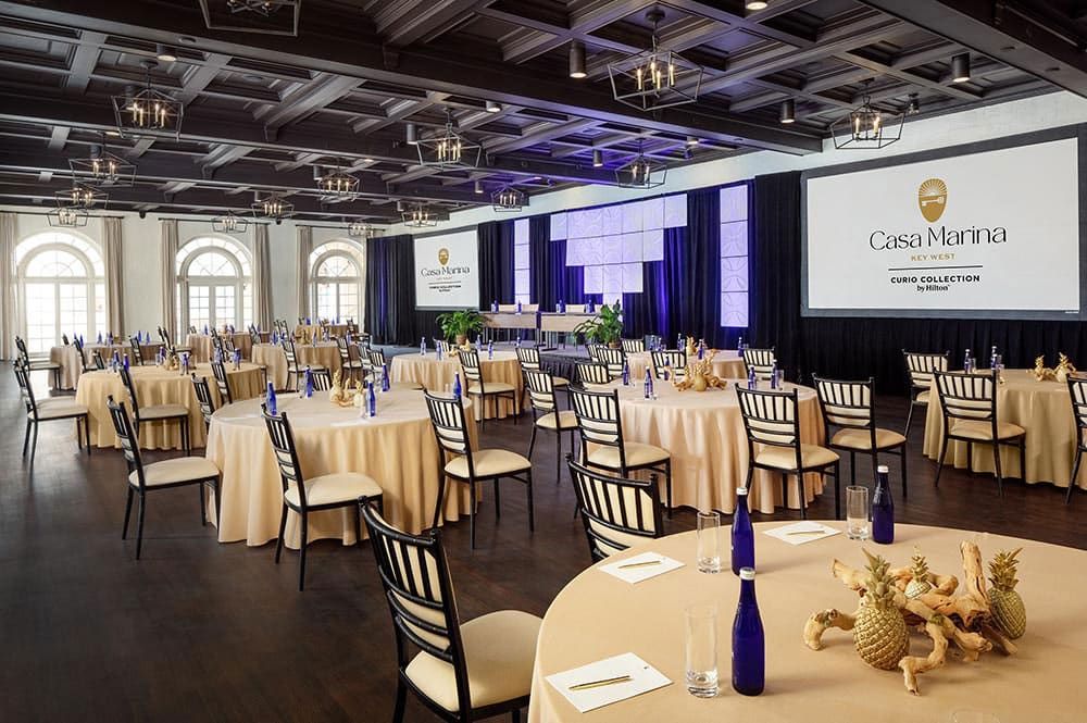 Spacious ballroom set for an event, with round tables, chairs, gold centerpieces, blue bottles, and projection screens displaying "Casa Marina Key West Curio Collection."