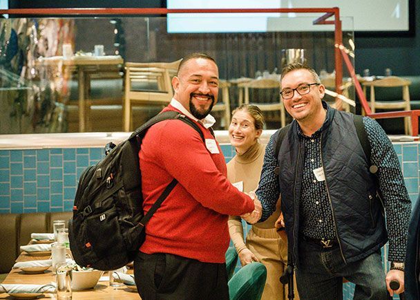 Three people are at a Retreats Resources roadshow event, with two men in the foreground shaking hands and a woman smiling in the background. Tables are set for dining.