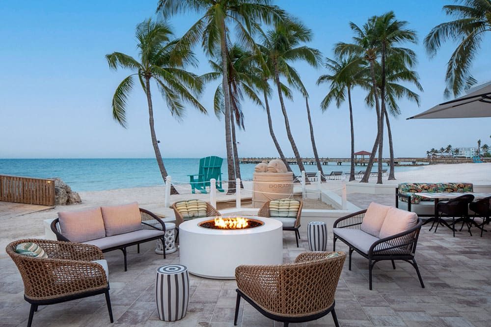 Outdoor seating area with wicker chairs and a fire pit on a patio overlooking a sandy beach, palm trees, and the ocean.